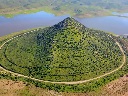 El **cerro Masatrigo**, cubierto de vegetación, se alza majestuoso junto a un embalse. Una carretera serpentea a su alrededor, conectando con un puente.