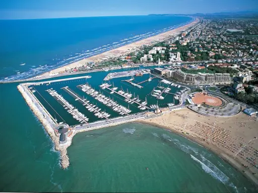 La darsena di Rimini, con le sue barche ormeggiate, si estende lungo la costa adriatica, tra la spiaggia affollata e il mare azzurro.