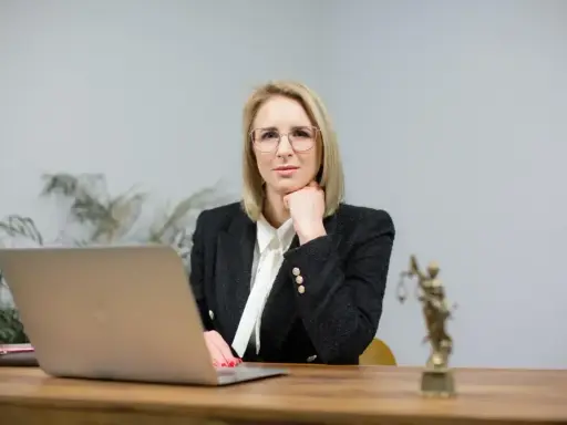Kobieta w okularach i żakiecie siedzi przy biurku z laptopem i Statuą Temidy.