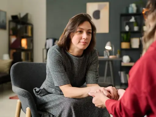 Kobieta w szarej sukience siedzi w fotelu, trzymając dłonie drugiej osoby. To scena wspierającej terapii gestalt.