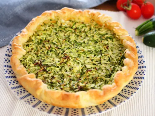 Torta salata con zucchine grattugiate e ricotta, con bordo dorato e decorazione blu su piatto bianco.