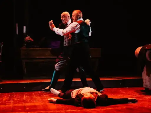 Dwóch mężczyzn tańczy tango na scenie, trzeci leży na podłodze.