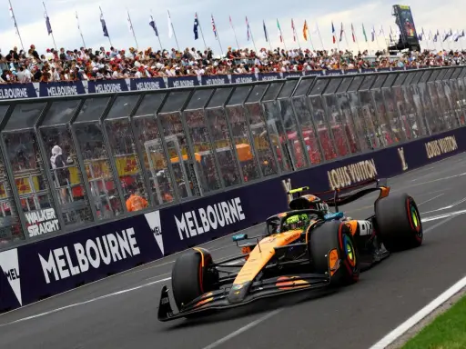 Un coche de Fórmula 1 naranja y negro acelera en Melbourne. Los aficionados vitorean. ¡Qué ganas de que empiece la Fórmula 1 2026!