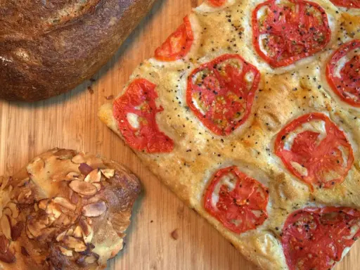 Focaccia receta con tomates frescos y hierbas, junto a un pan rústico y un bollo con almendras.