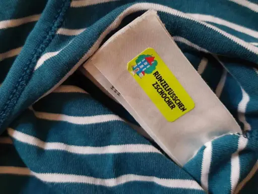 Namensetiketten Kita Erfahrungen: Ein gestreiftes Shirt mit einem gelben Aufkleber "Runzelfüßchen Zschocher" und einem Hausmotiv.