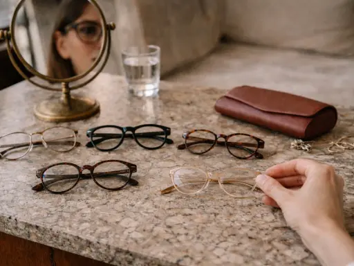 Kobieta z twarzą diamentową wybiera okulary. Na stole leżą różne modele, portfel i biżuteria.