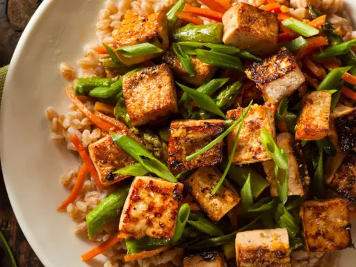 Talerz pełen brązowego ryżu z grillowanym tofu, szparagami i marchewką, posypany szczypiorkiem.