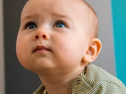 Occhi azzurri di un neonato di 10 mesi che guarda in alto, con un'espressione curiosa. Indossa una maglietta a righe gialle e nere.