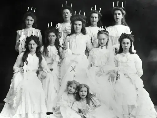 En grupp flickor i vita klänningar, några med ljus i håret, visar hur Lucia såg ut förr.