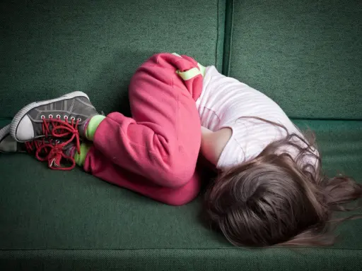 Dziecko skulone na kanapie, w różowych spodniach i bluzce w paski. Widać buty i skarpetki.