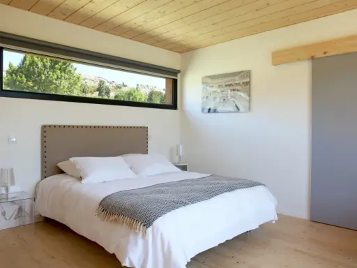 Chambre couleur taupe avec un grand lit blanc, une tête de lit capitonnée et une vue sur la nature par la fenêtre.