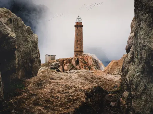 Leuchtturm auf felsiger Küste der Costa da Morte, Vögel ziehen über neblige Berge.