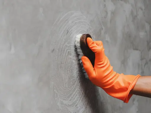 Mano con guante naranja limpiando pared gris con cepillo, aprendiendo como limpiar las paredes de color sin dañar la pintura.