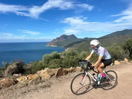 Ciclista pedala lungo la costa rocciosa della Corsica, con il mare blu e montagne sullo sfondo. Un'avventura in bici indimenticabile.
