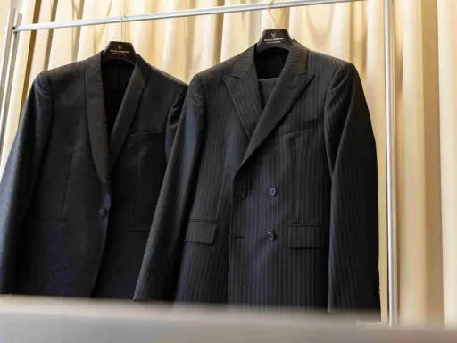 Dwa eleganckie garnitury wiszą na wieszakach. Jeden w prążki, drugi z subtelnym wzorem. Prasowanie garnituru to klucz do nienagannego wyglądu.