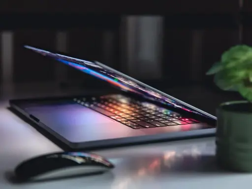 Otwarty notebook z podświetlaną klawiaturą i ekranem, obok myszka i roślina. To jest właśnie notebook, narzędzie do pracy i rozrywki.