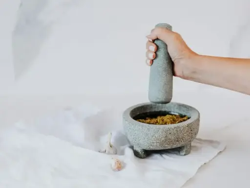 Dłoń uciera w moździerzu aromatyczne pesto z bazylii. Obok leżą ząbki czosnku.
