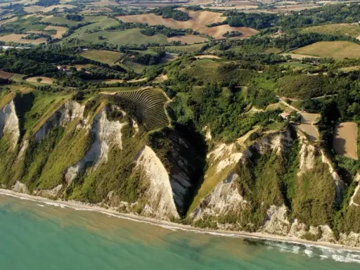 Scogliere a picco sul mare turchese a Gabicce Mare, Marche. Campi coltivati e vegetazione rigogliosa adornano le colline retrostanti.
