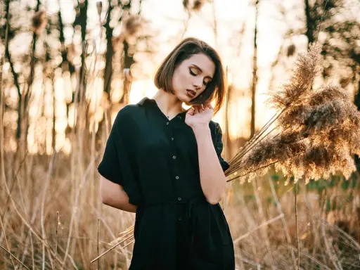 Młoda kobieta w czarnej sukience pozuje na tle zachodzącego słońca i trzcin. Idealna modelka do nauki kurs fotografii portretowej.