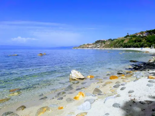 Spiaggia di Tropea con la torre Sant'Irene in lontananza, mare cristallino e rocce sulla riva.