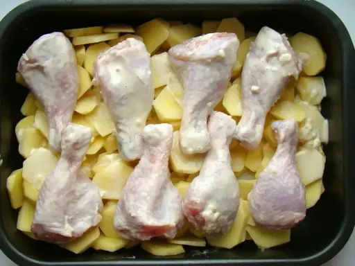 Osiem pałek kurczaka w sosie, ułożonych na plastrach ziemniaków w brytfannie. Idealne do pieczenia, by uzyskać chrupiące pałki z kurczaka w piekarniku.