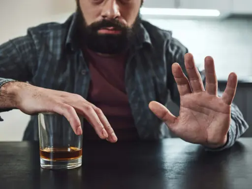 Mężczyzna z zarostem odpycha szklankę z alkoholem, walcząc z chorobą alkoholową. Gest dłoni symbolizuje odmowę i chęć zmiany.