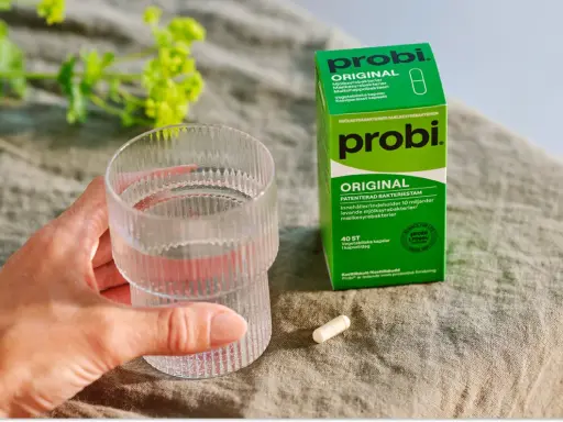 En hand håller ett glas vatten bredvid en förpackning med probi. En kapsel ligger på bordet, redo att tas för att undvika probi mage biverkningar.