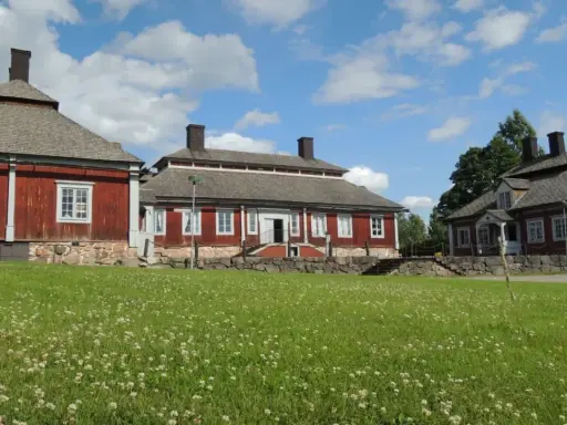 Malingsbo herrgård med sina röda trähus och stenfundament, omgivet av en grönskande äng med vita blommor under en blå himmel med moln.