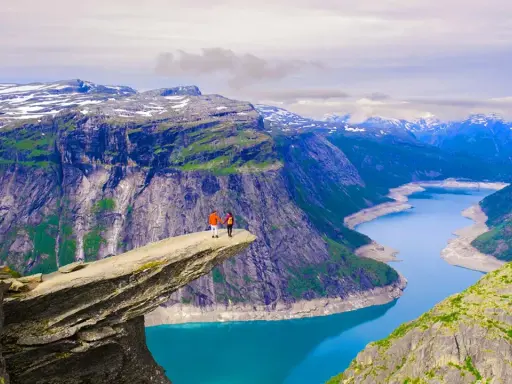 Två personer står på Trolltunga, en av Norges mest ikoniska och fina platser. En dramatisk klippavsats med en turkos sjö nedanför och snöklädda berg i bakgrunden.
