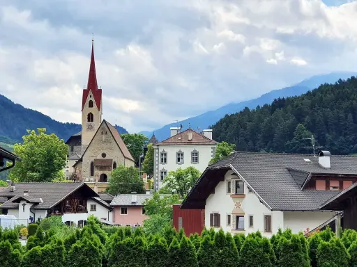 Un borgo pittoresco in Val d'Isarco, con una chiesa dal campanile rosso svettante tra case tradizionali e montagne boscose.