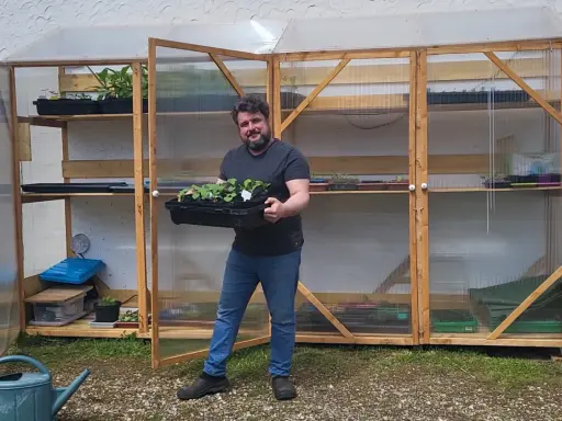 Un homme souriant tient un plateau de jeunes plants devant sa serre pour semis fait maison, remplie d'étagères de semis.