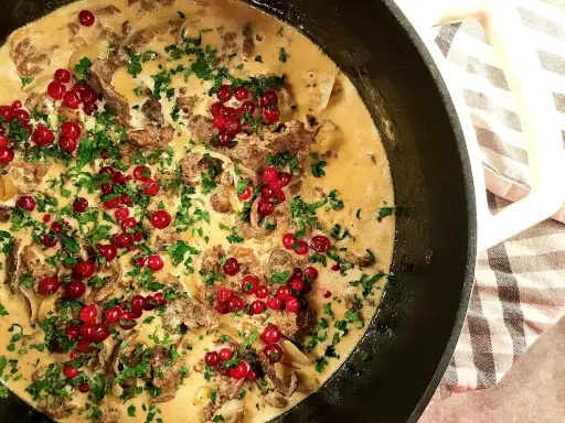 En närbild på en krämig renskavsgryta med färska lingon och persilja, serverad i en gjutjärnspanna.