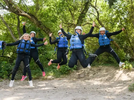Cinco personas, incluyendo niños, saltan alegres con trajes de neopreno y chalecos salvavidas en un entorno natural, listos para una aventura en Huesca.