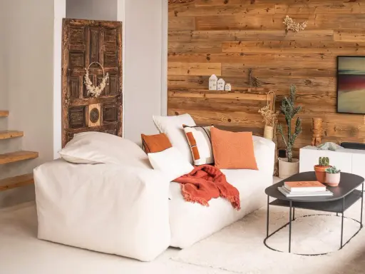 Un salon chaleureux et accueillant avec un grand canapé blanc, des coussins orange et un mur en bois rustique. Une porte ancienne décore le mur.