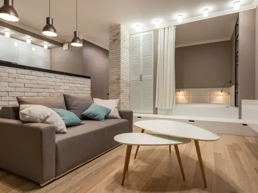 Nowoczesny salon z sofą i stolikami kawowymi. W tle widoczna ściana ze starej cegły, łóżko i szafa.