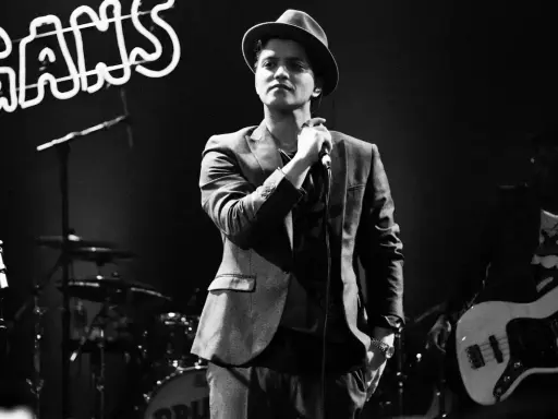 Bruno Mars i Sverige, med hatt och kavaj, sjunger i mikrofonen. Bakom honom syns ett band och neonljus.