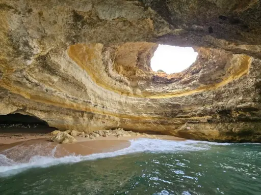 Die Benagil-Höhle mit ihrem kreisförmigen Loch im Felsdach, das Sonnenlicht hereinlässt. Wellen brechen sanft am Sandstrand im Inneren.