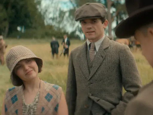 Giovane donna con cappello a scacchi e gilet a rombi, accanto a un uomo con berretto e giacca di tweed. Sullo sfondo, i sette quadranti di un campo.