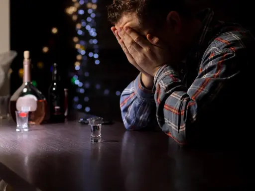 Mężczyzna z rękami na twarzy przy barze, obok butelka i kieliszek. Obraz symbolizuje przymusowe leczenie alkoholika i jego walkę.