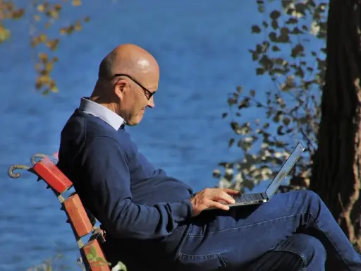 Mężczyzna w okularach siedzi na ławce nad jeziorem, pracując na laptopie. Planuje swoje ubezpieczenie emerytalne, ciesząc się spokojem natury.