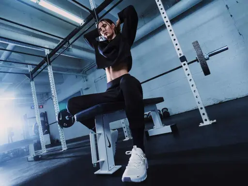 Donna in tenuta sportiva nera, con scarpe bianche, seduta su panca in palestra. Ispirazione su come vestirsi in palestra.