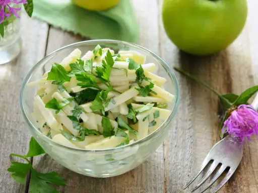 Frischer Kohlrabi Salat mit Petersilie in einer Glasschüssel, garniert mit einer lila Blume und grünen Äpfeln auf Holztisch.