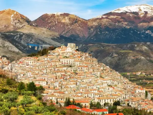 Un pittoresco borgo calabrese si aggrappa a una collina, con case in pietra e tetti rossi che si estendono fino alle montagne innevate.