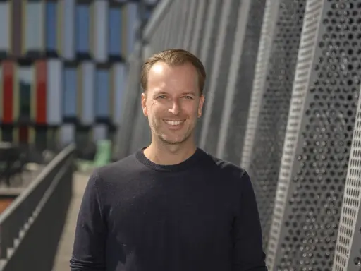 Daniel Larsson, skådespelare, ler mot kameran. Bakgrunden är en modern byggnad med geometriska mönster.