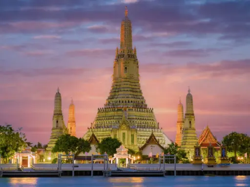Il **tempio dell'alba Bangkok** (Wat Arun) si erge maestoso al tramonto, con le sue guglie dorate che brillano contro un cielo rosa e viola.