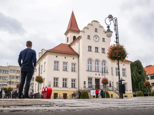 Mężczyzna podziwia ratusz, jedną z głównych trzebnickich atrakcji. W tle czerwone serce i kwitnące drzewka.