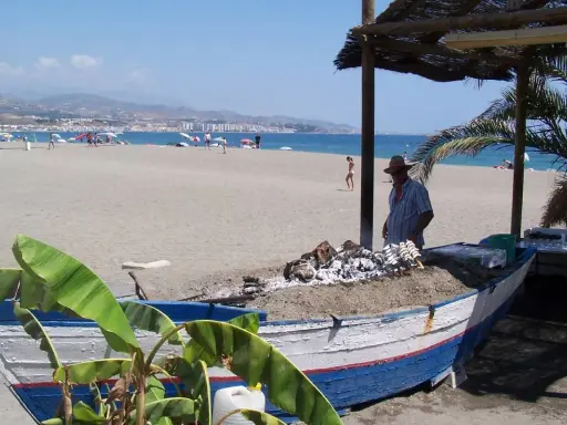 Ein Mann grillt Fisch auf einem Strand in Torre del Mar. Im Hintergrund das Meer und Berge.