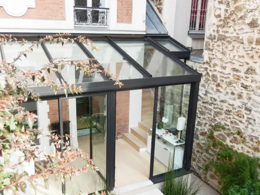 Une véranda bioclimatique moderne, avec une structure en verre et métal noir, s'intègre à une maison en briques et pierre. Avis positif sur son esthétique.