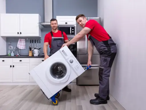 Dwóch mężczyzn w roboczych ubraniach przewozi pralkę na wózku. Czy można przewozić pralkę na leżąco? To pytanie zadaje sobie wielu przy przeprowadzce.
