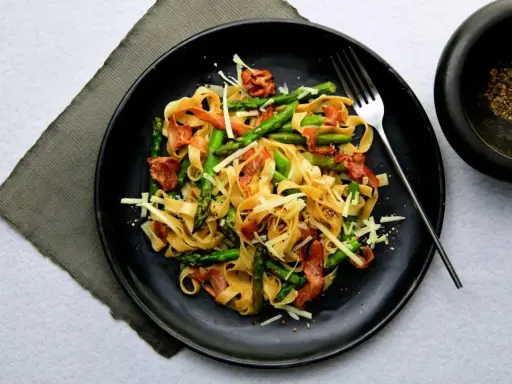 Pyszny makaron ze szparagami, chrupiącym boczkiem i parmezanem na czarnym talerzu. Idealny na wiosenny obiad.